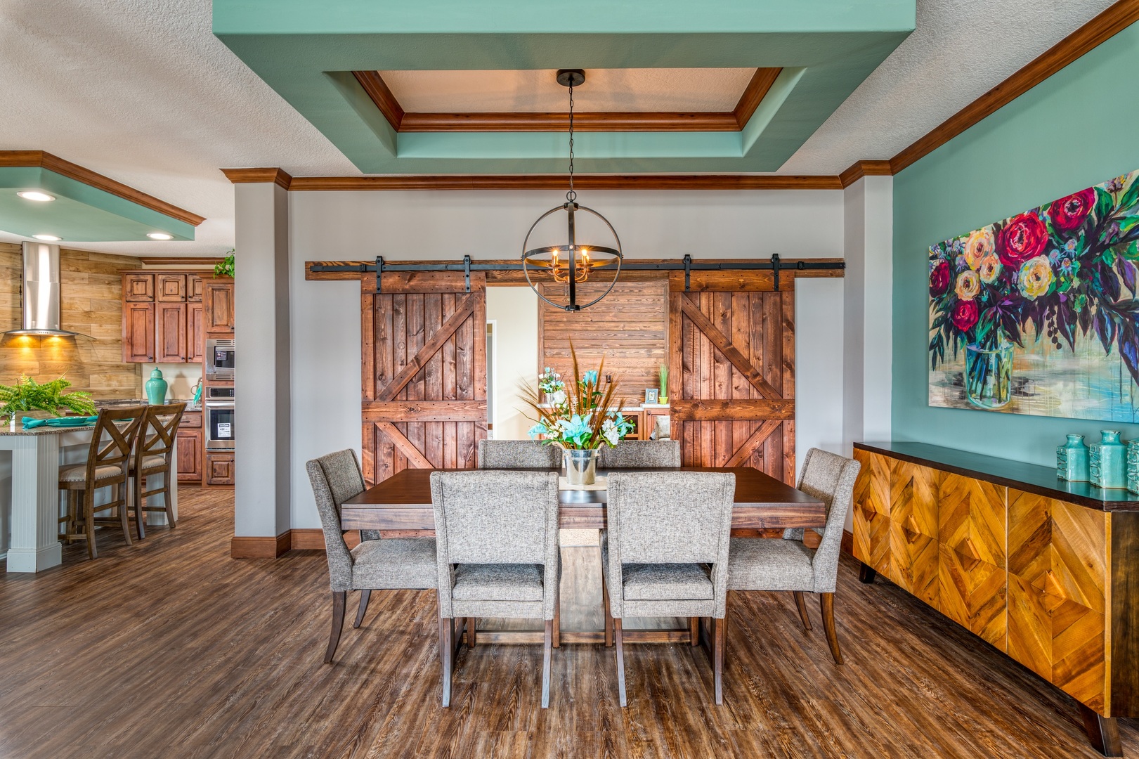 A modern dining room with a wooden table, six gray upholstered chairs, and a rustic sliding barn door. Vibrant floral artwork hangs above a sideboard.