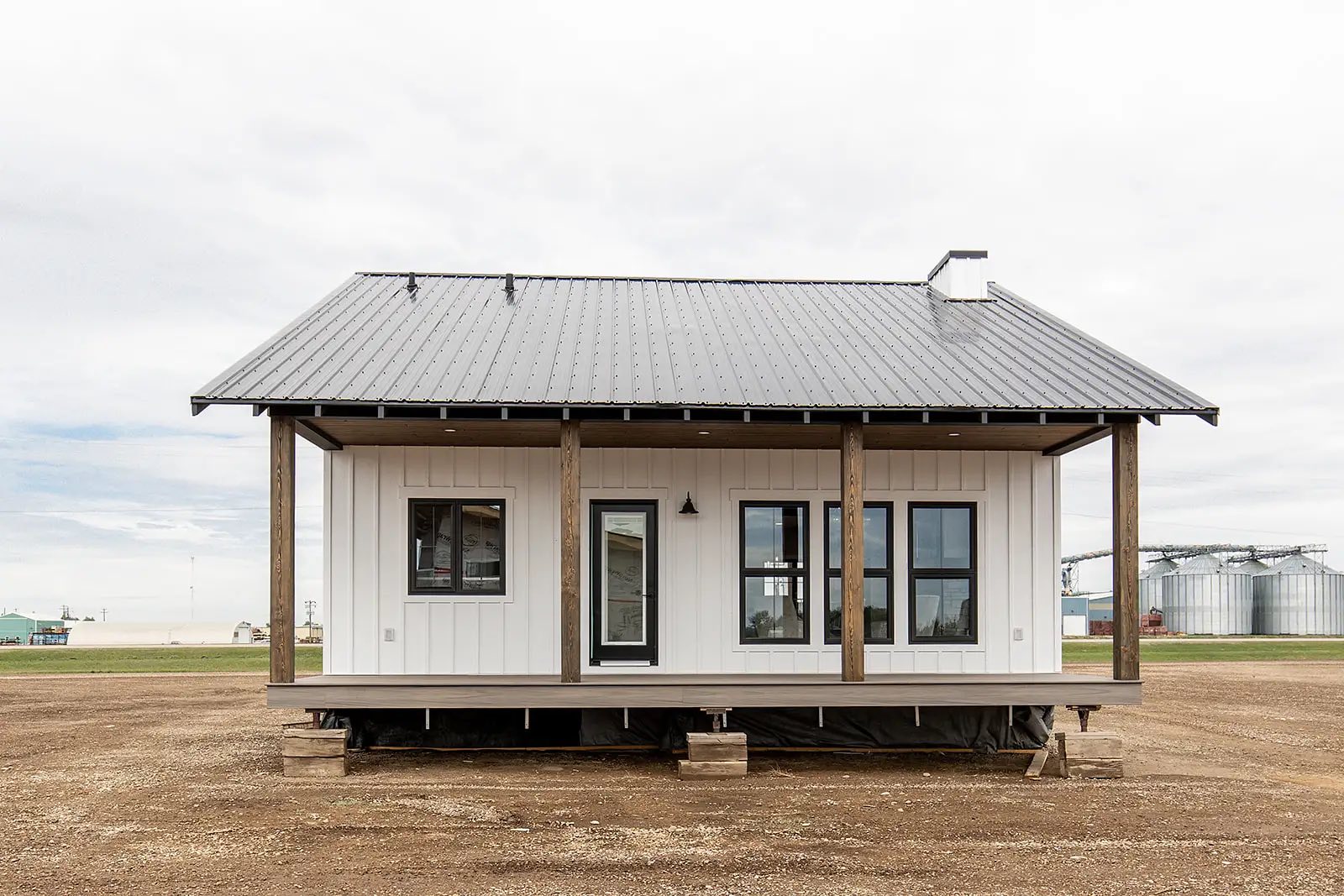 A small, white, one-story farmhouse with a metal roof and wooden columns on an elevated platform, set against a rural landscape with overcast skies.