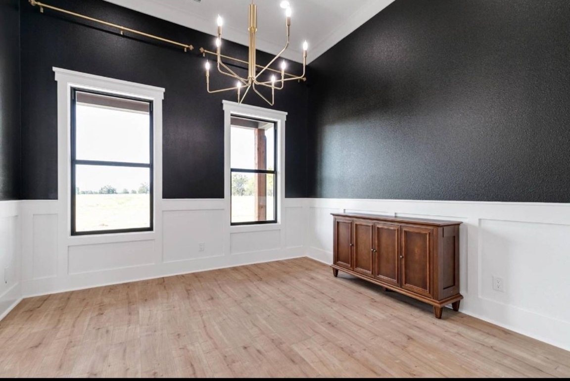A minimalist room with black walls and white wainscoting features two tall windows. A modern gold chandelier hangs above a wooden sideboard on light wood flooring.