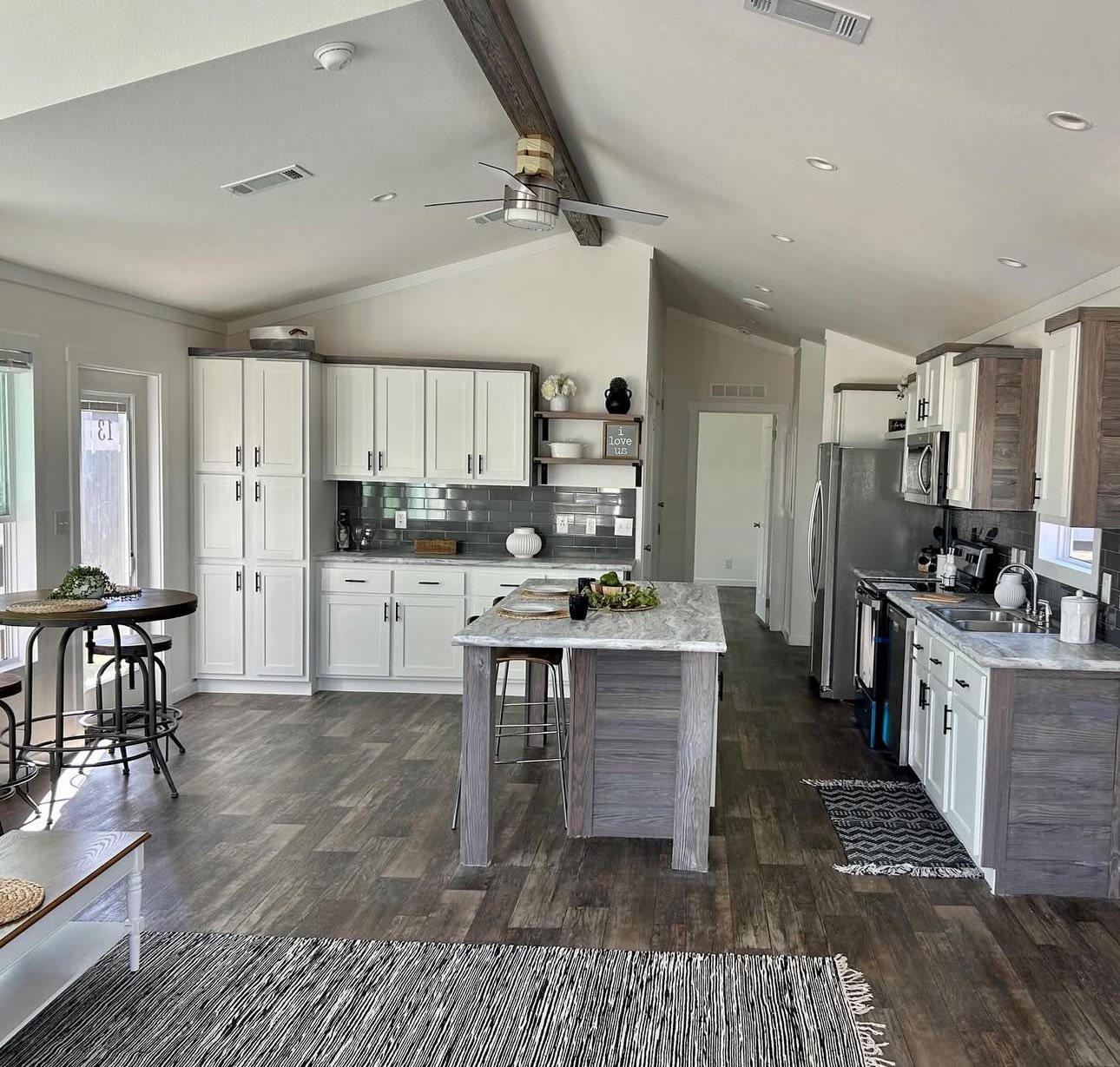 Spacious kitchen with high ceiling and dark wood beam, featuring white cabinets, gray backsplash, marble island, wooden floor, and cozy dining area.