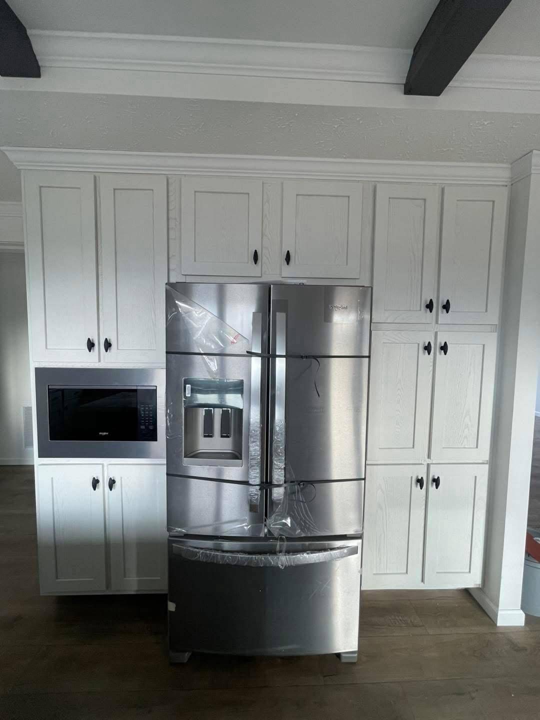 Stainless steel French door fridge in a modern kitchen, surrounded by white cabinetry and a built-in microwave. Shelving and beams add contrast.