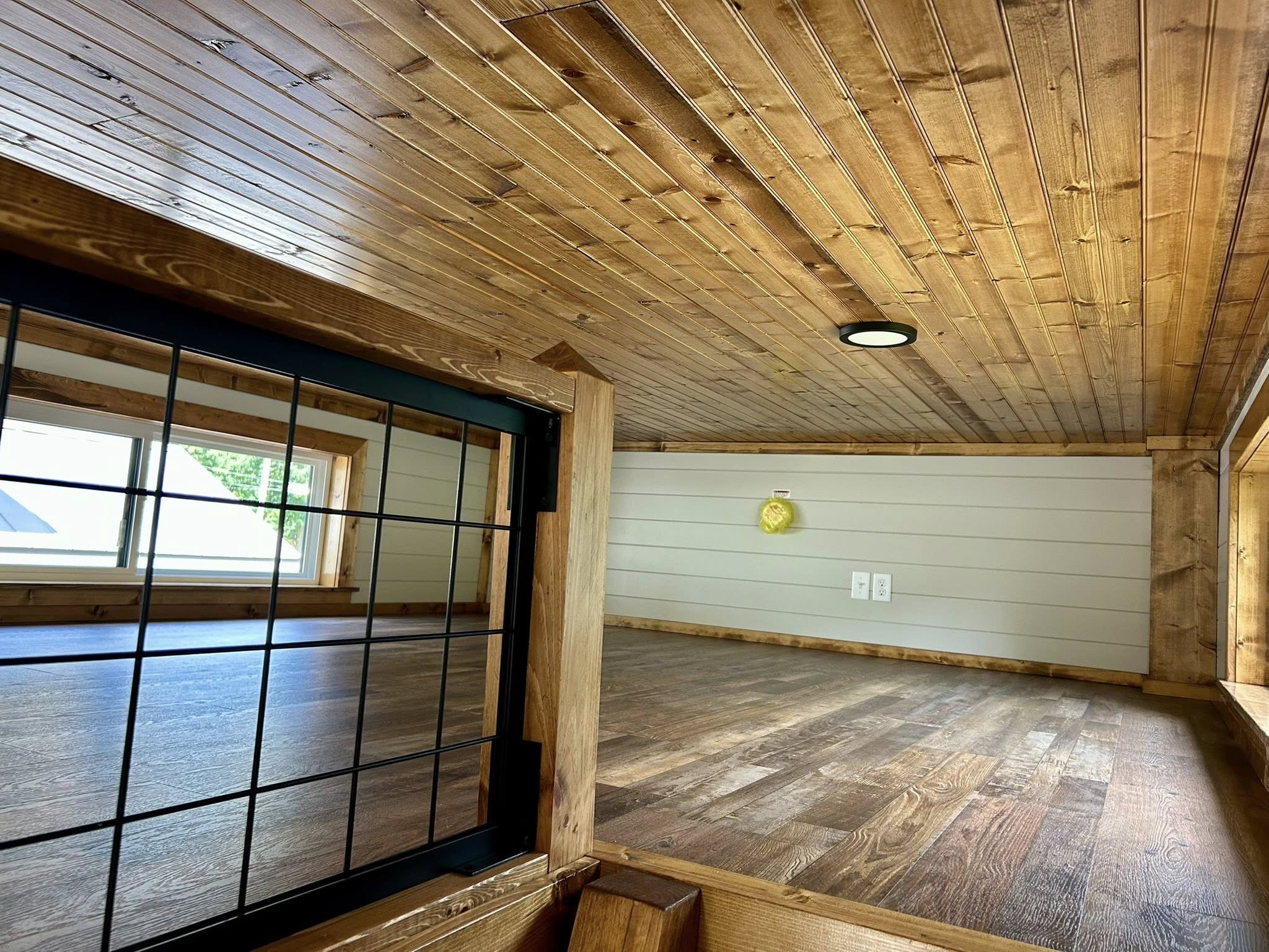 Cozy, rustic loft with wooden ceilings and floors, large windows with grid details, light walls, and a small yellow clock. Inviting and serene space.