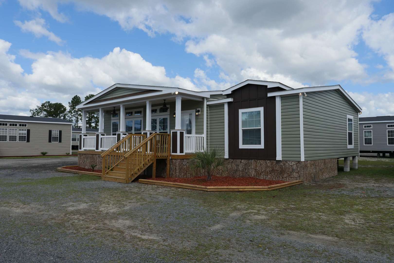 A modern manufactured home with a green exterior, white trim, and stone foundation. It features a wooden porch with stairs, set under a cloudy sky.