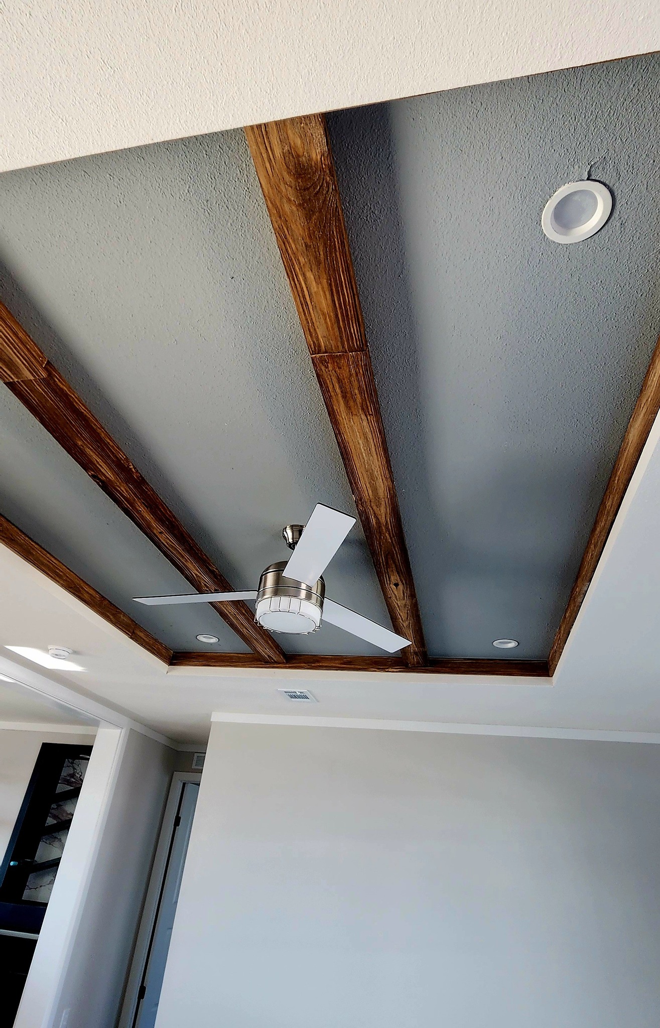 Ceiling with wooden beams and modern white fan, surrounded by recessed lights. The grey contrasts with the wood, creating a stylish look.