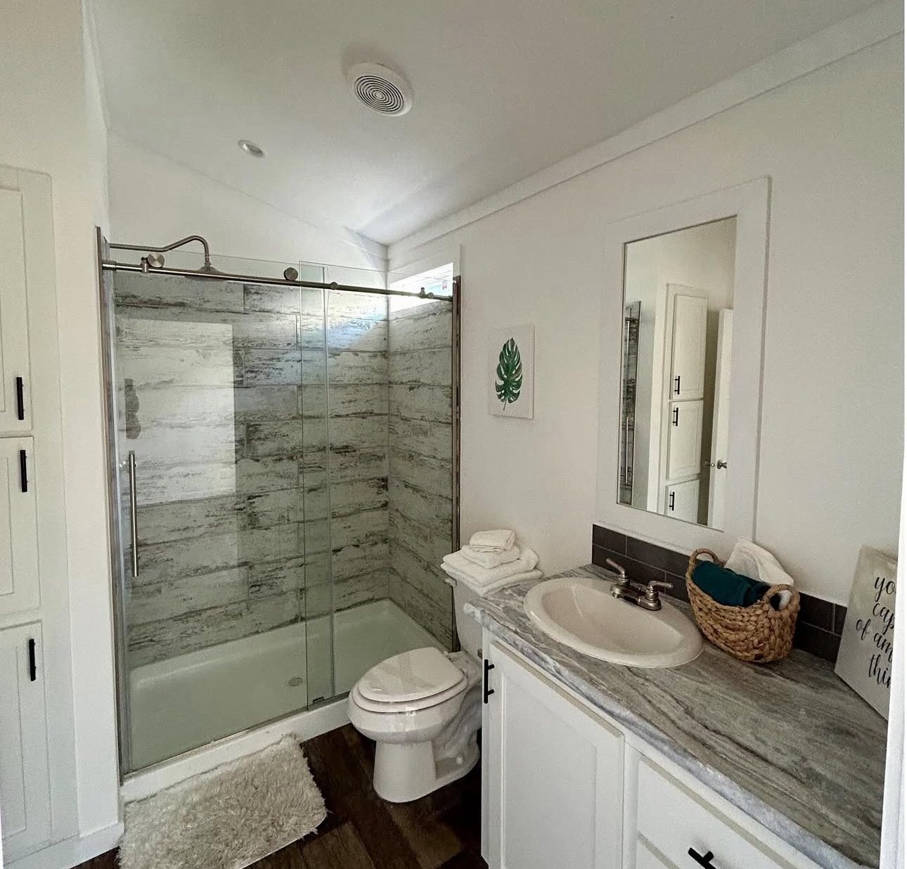 Modern bathroom with a glass-enclosed shower, marble walls, and wood flooring. Features a white vanity, a basket of towels, and leafy wall art.