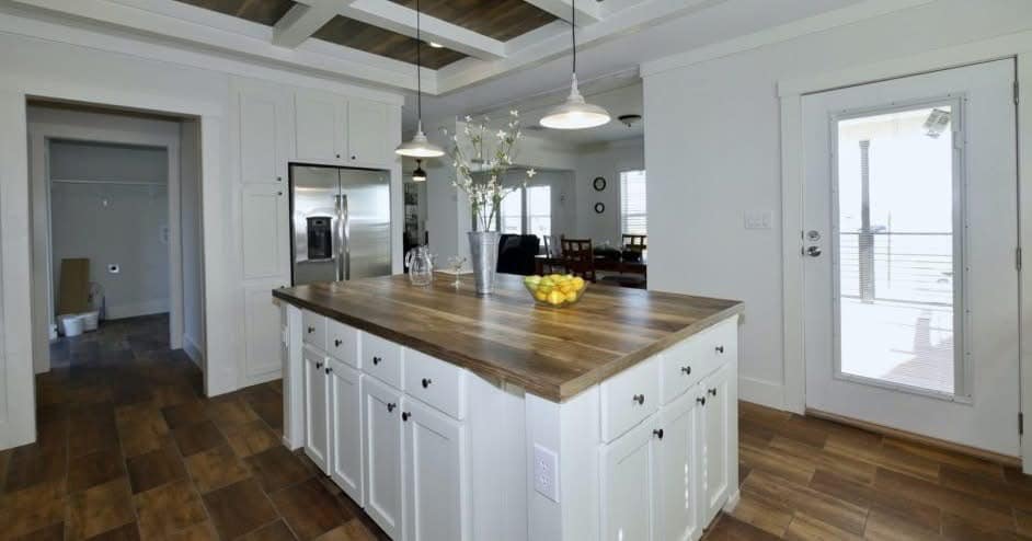 Spacious modern kitchen with white cabinetry, a wooden island countertop holding vases and fruit, pendant lights, and a sunlit door. Cozy, welcoming ambiance.