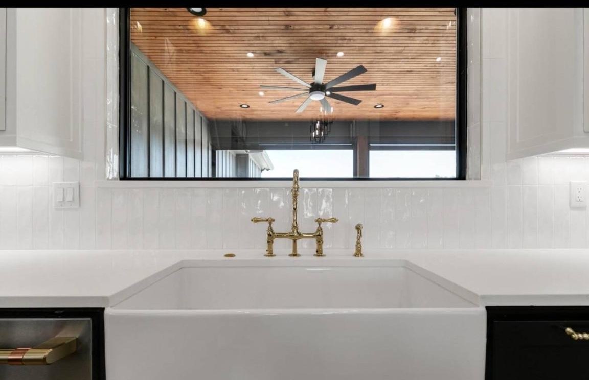 A white farmhouse sink with brass faucets is set against a bright kitchen. A large window overlooks a wooden patio ceiling with a modern black fan.