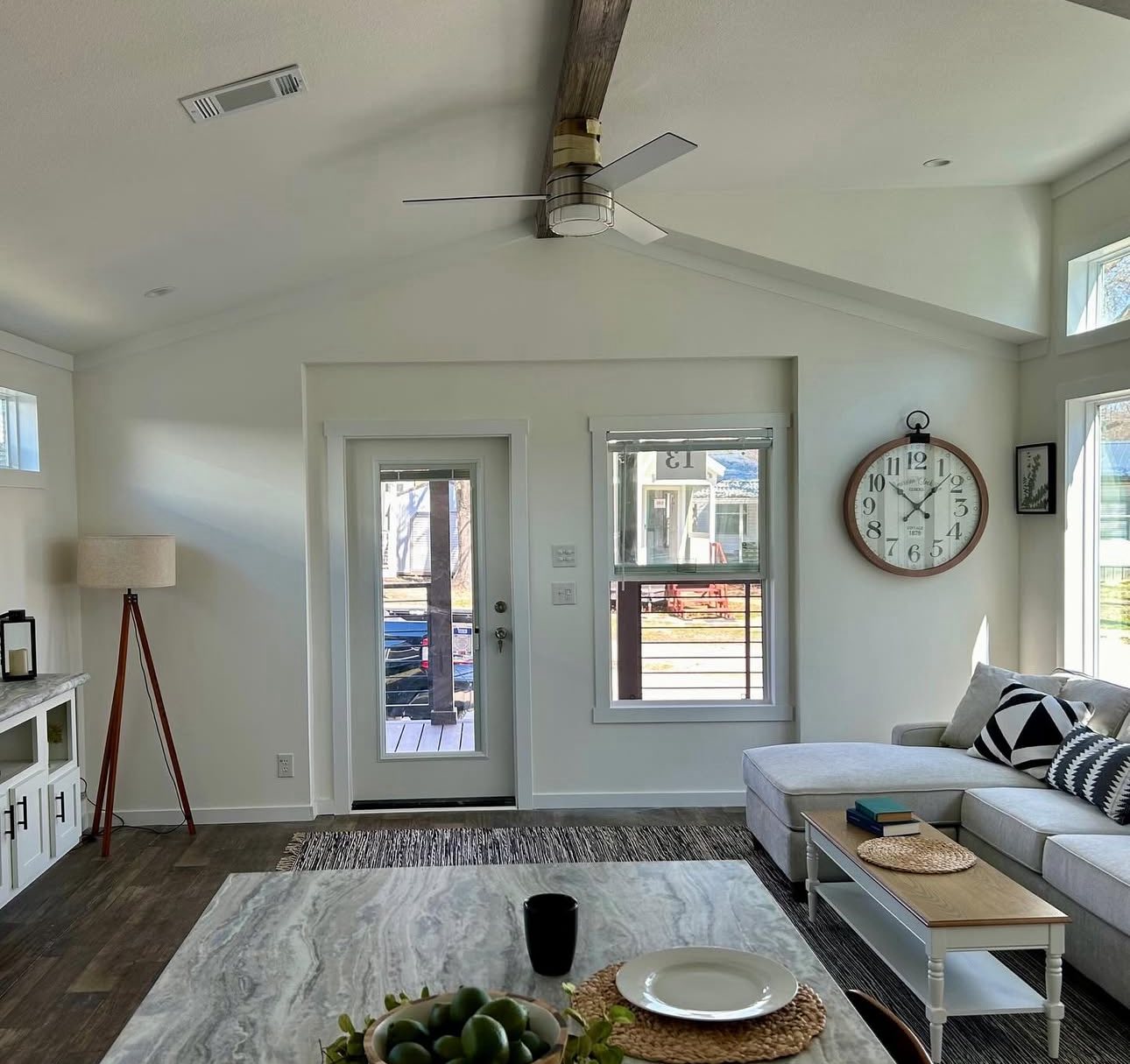 Bright living room with a white interior, featuring a large wall clock, beige couch, patterned pillows, and a marble coffee table on a rug. A door and window let in natural light, creating a cozy, modern atmosphere.