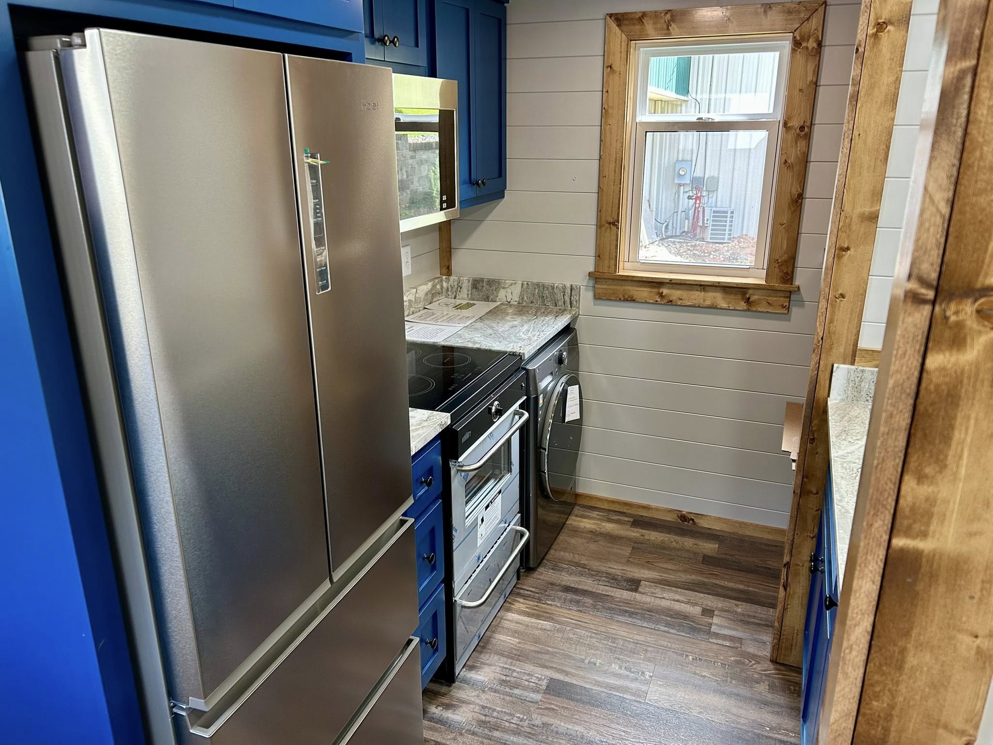 Compact kitchen with wood floors, blue cabinets, and modern appliances. A stainless steel fridge and oven are next to a window framed in wood.