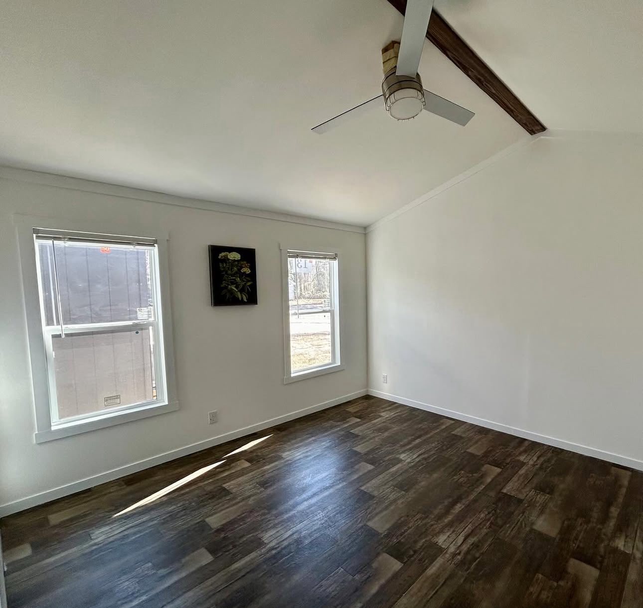 A bright, empty room features dark wood floors, two windows with natural light, a ceiling fan, and a floral painting on a white wall, creating a clean, modern feel.