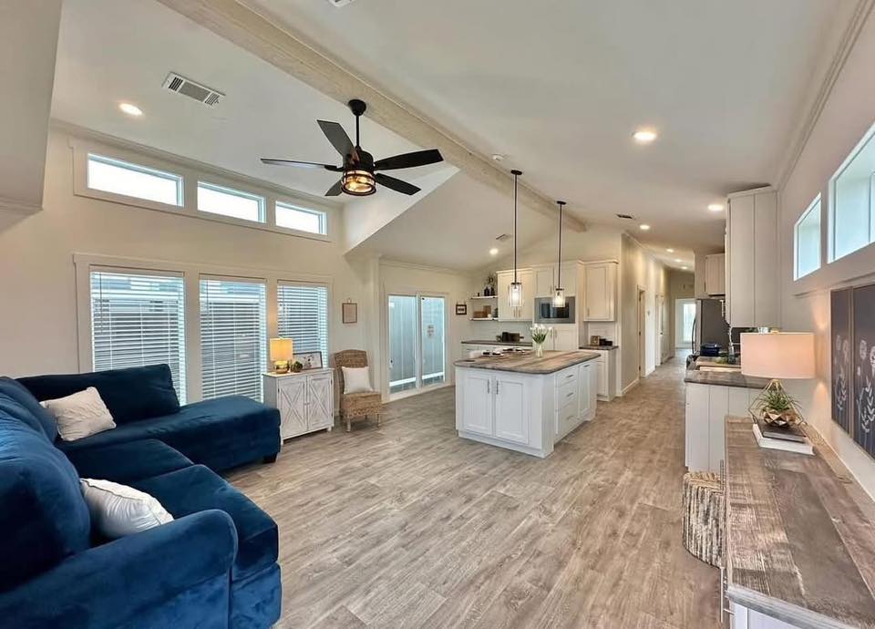 Spacious open-plan living room and kitchen with light wood flooring. Features a navy sofa, modern ceiling fan, a central island, and minimalist decor. Bright and inviting atmosphere.