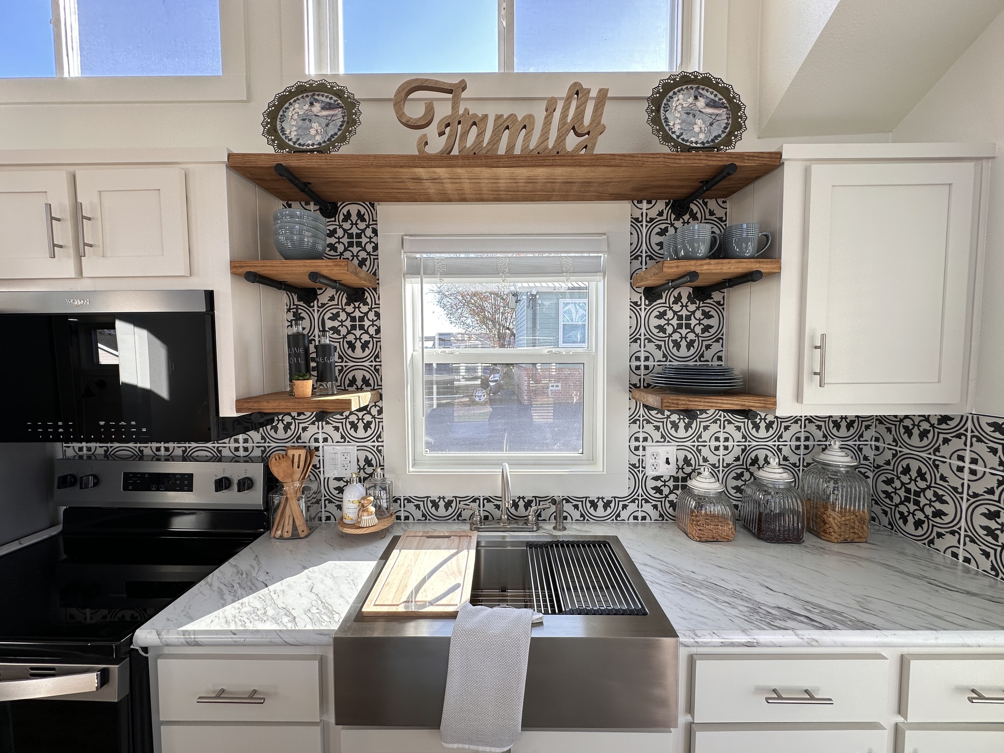 A cozy kitchen features a marble countertop, farmhouse sink, patterned backsplash, and shelves with jars, dishes, and a "Family" sign above. Bright, inviting feel.