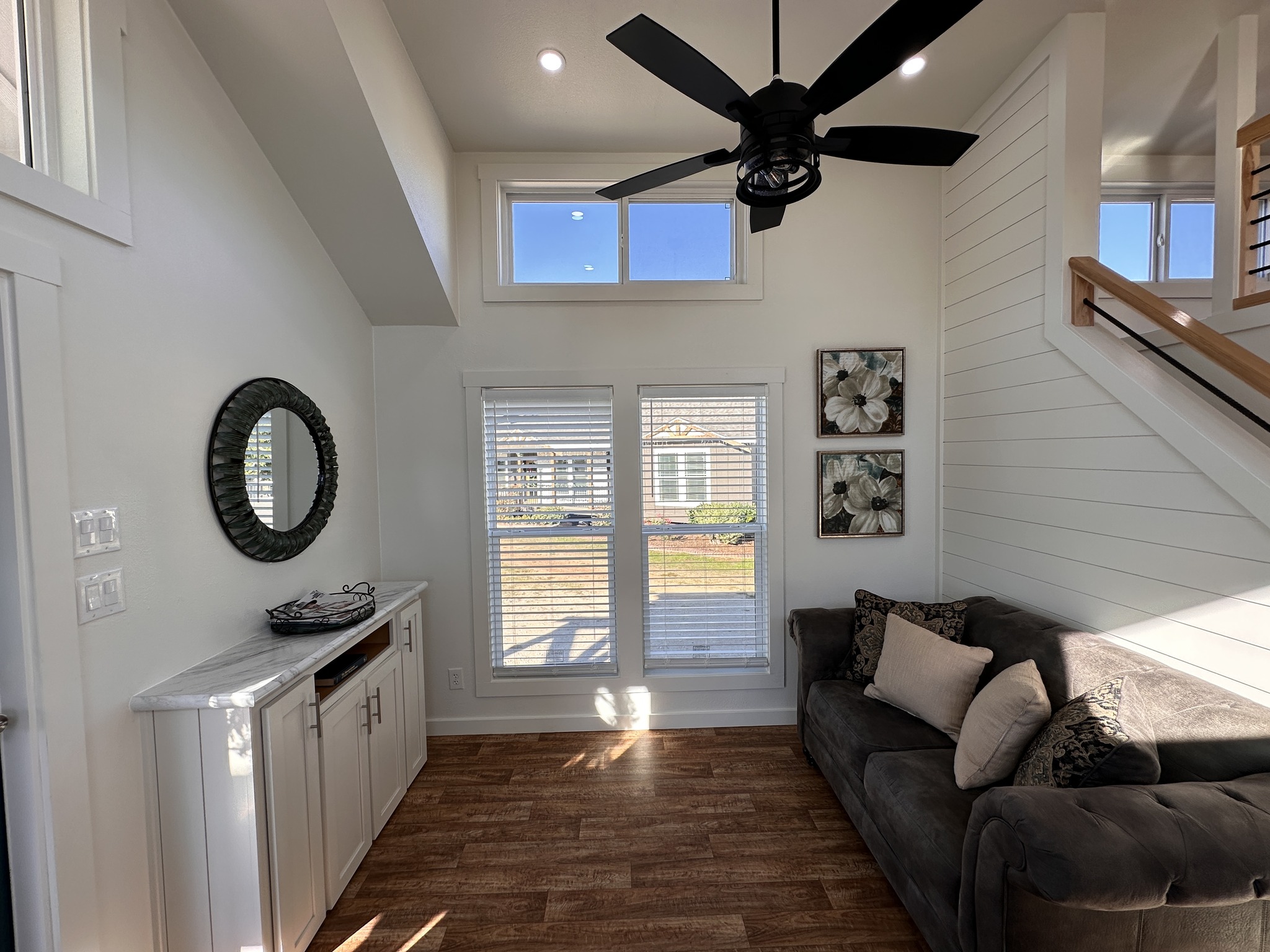 Bright living room with large windows, light wood floors, a gray sofa with cushions, and wall art. Black ceiling fan adds contrast. Cozy and inviting.