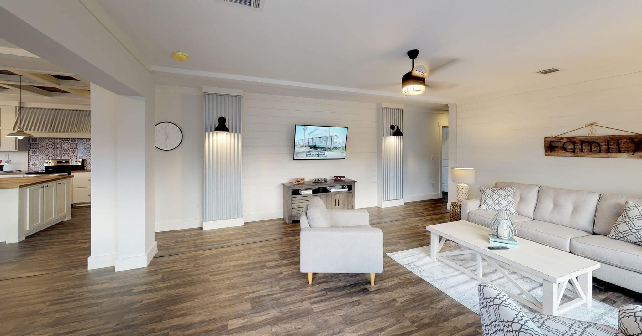 Spacious living room with a beige sofa, patterned pillows, and a white coffee table on a rug. Wall-mounted TV, wooden floors, and a rustic "Family" sign. Bright and inviting atmosphere.