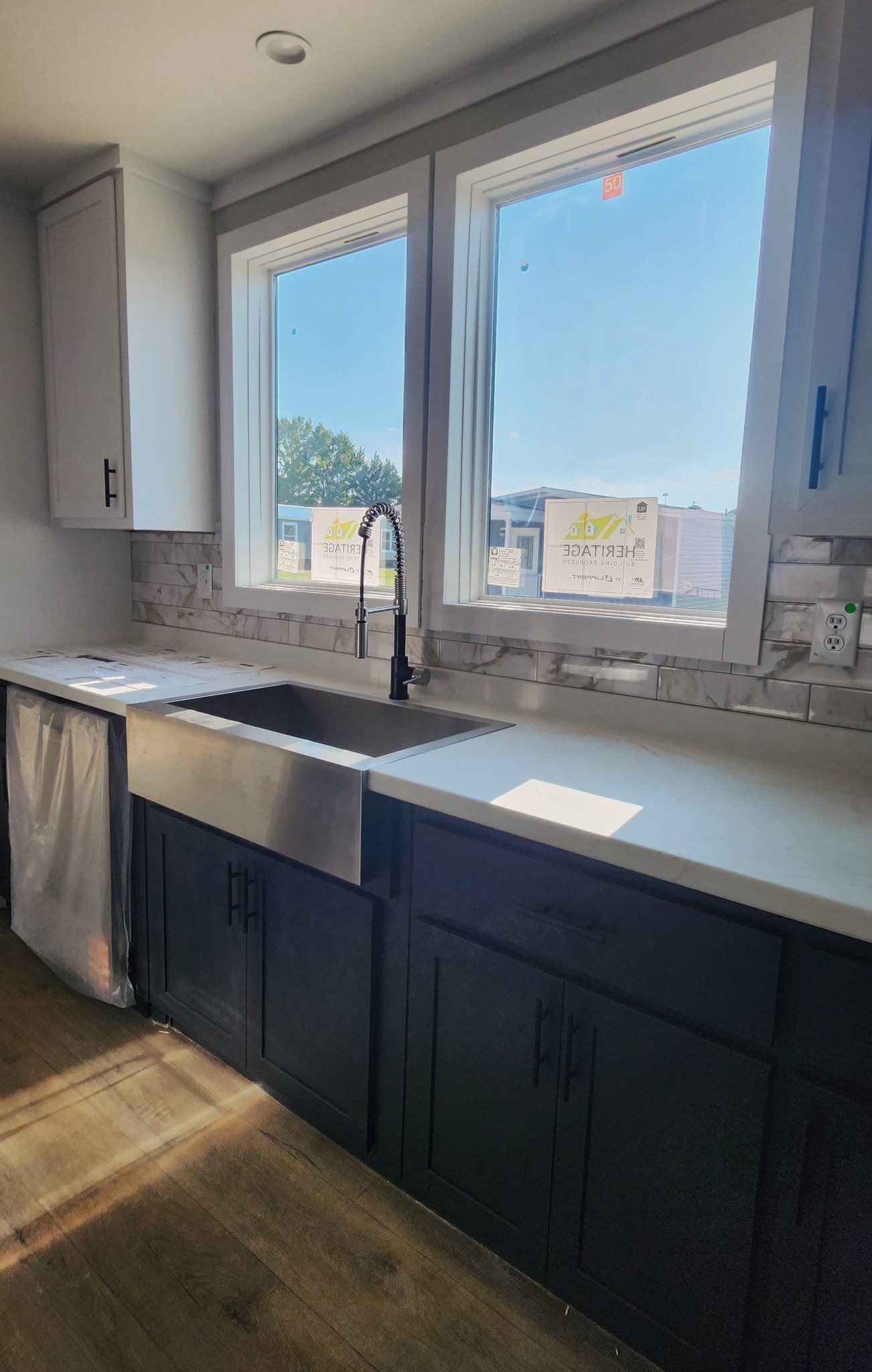 Bright kitchen with dual windows above a stainless steel sink and dark cabinetry. Sunlight casts patterns on the wooden floor, creating a warm, inviting tone.