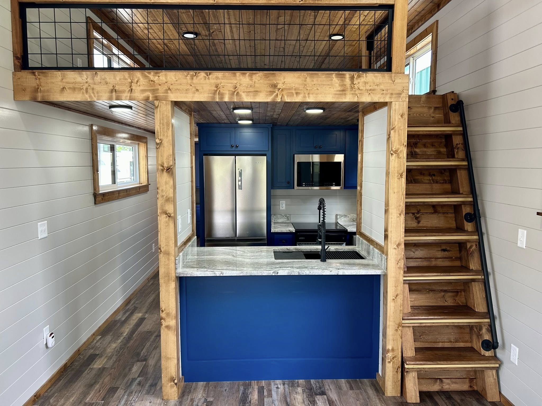 Compact kitchen with blue cabinets, stainless steel appliances, and a marble countertop. Wooden stairs lead to a loft, with cozy, modern vibe.