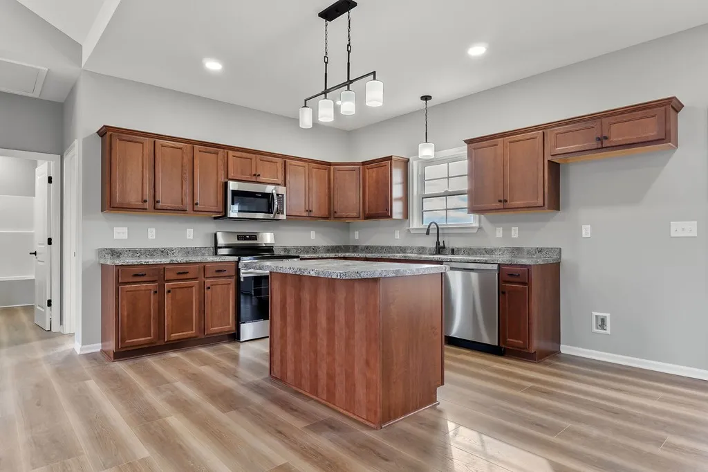 Spacious kitchen with wood cabinets, granite countertops, and stainless steel appliances. A central island under modern hanging lights on hardwood floors.