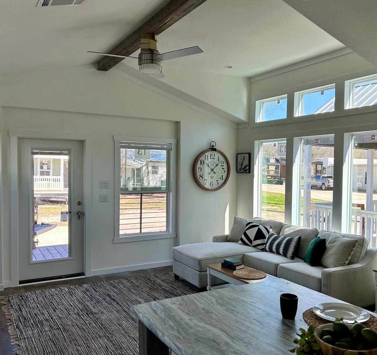 Bright living room with large windows and vaulted ceiling, featuring a cozy beige sectional adorned with modern pillows, a round wall clock, fan, and minimalist decor. A textured rug rests on dark wood flooring, adding warmth to the airy space.