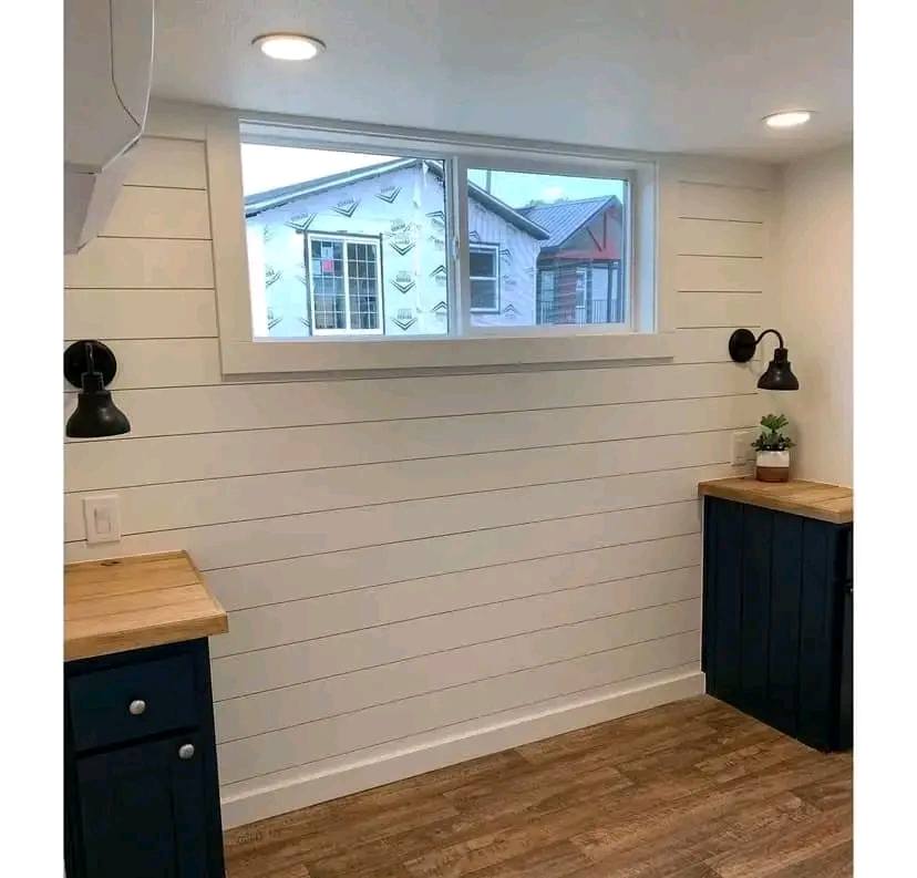Cozy room with white shiplap walls, wooden countertops, blue cabinets, and black sconces. Bright window views outside; hardwood flooring adds warmth.
