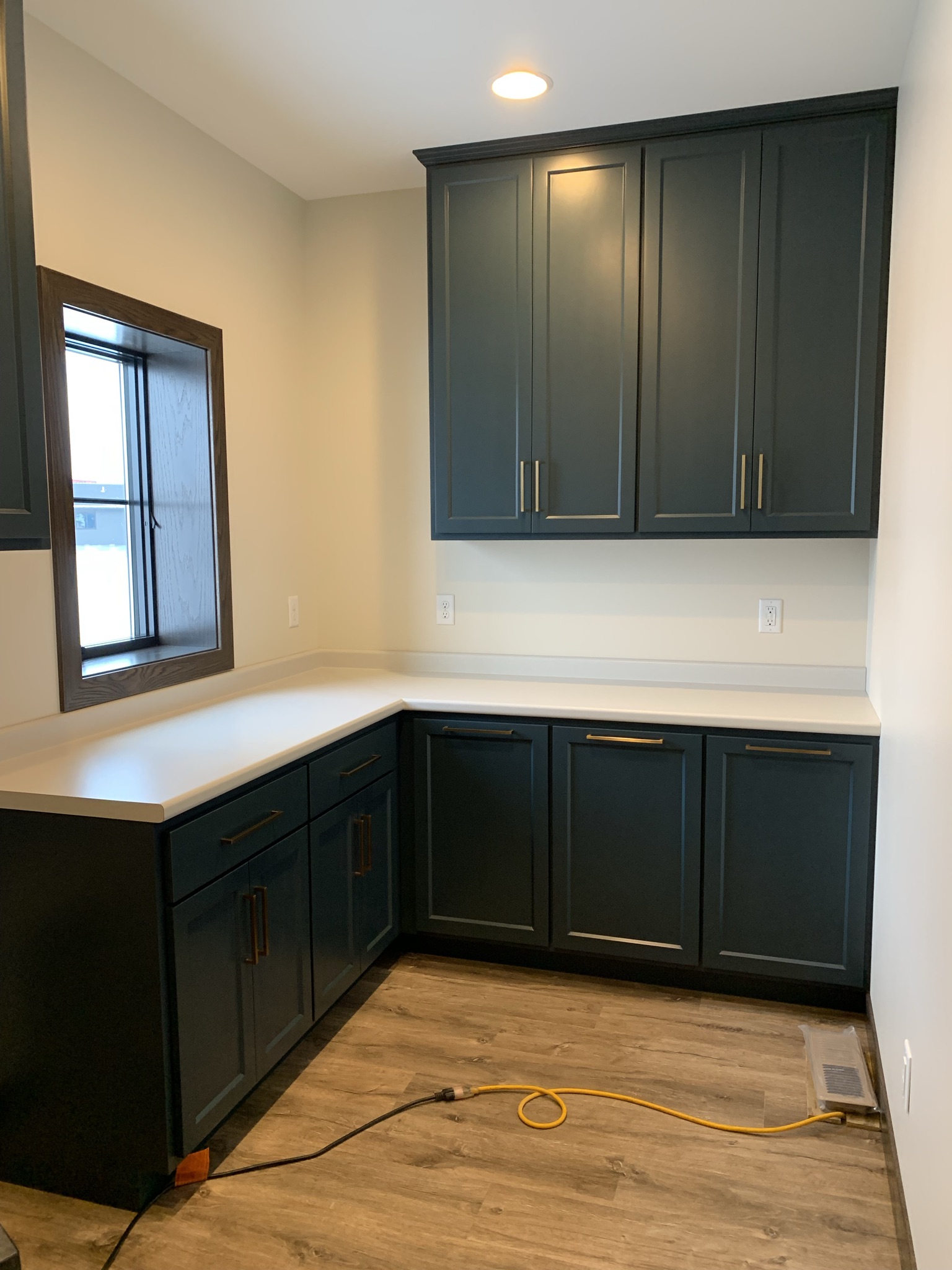 Compact kitchen with dark blue cabinets, sleek brass handles, and light countertops. A lit recessed light adds warmth; a coiled yellow cord on the wood floor.