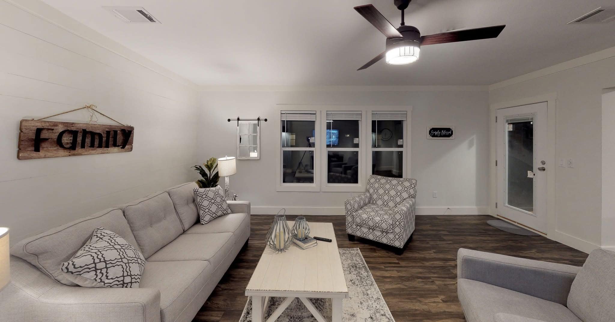 A cozy living room with a gray sofa and two armchairs, wooden coffee table, ceiling fan, family sign, and large windows. Warm and welcoming atmosphere.