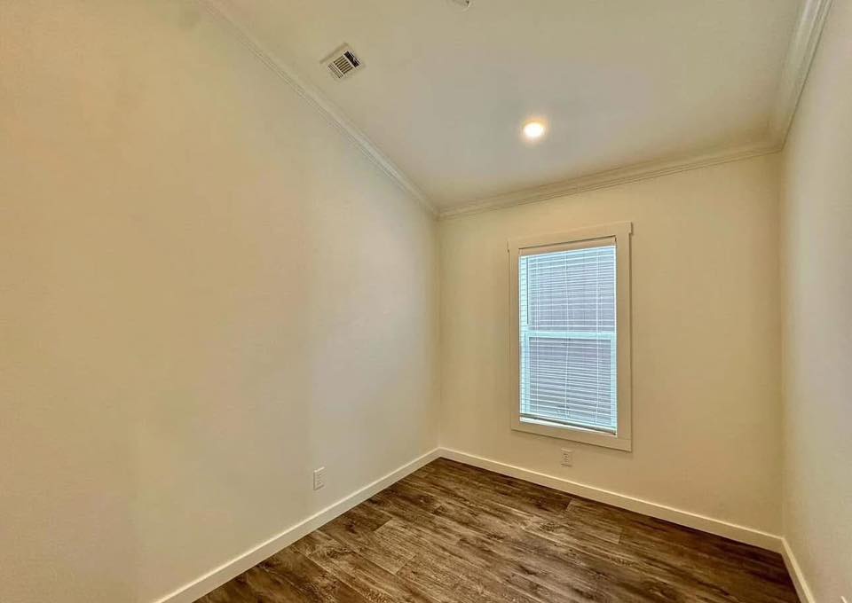 Small, empty room with cream walls and wood flooring. A single window with blinds lets in natural light from the right, illuminating the cozy space.