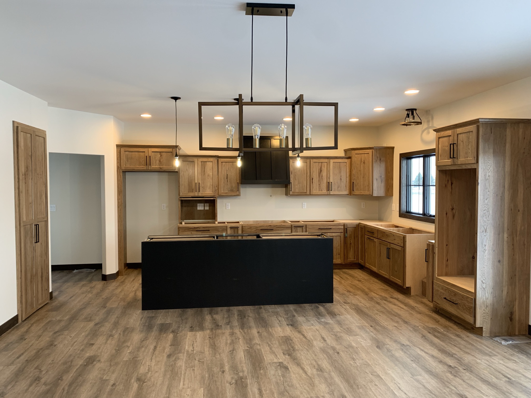 Spacious kitchen with wooden cabinets and dark accents. Features a central black island, pendant lights, and large window. Rustic and modern feel.