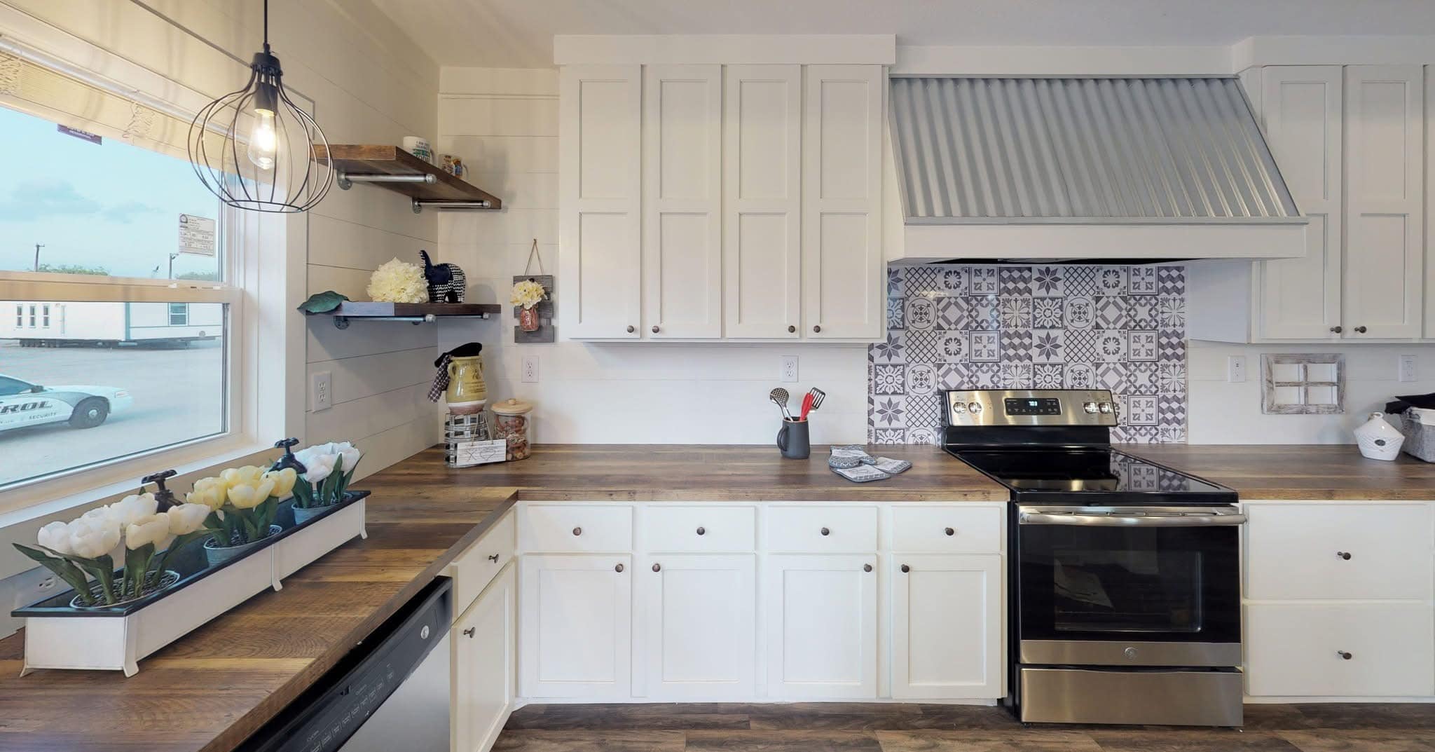 Bright kitchen with white cabinets, wood countertops, and a patterned backsplash. A sleek stainless steel oven sits beneath a corrugated metal range hood.
