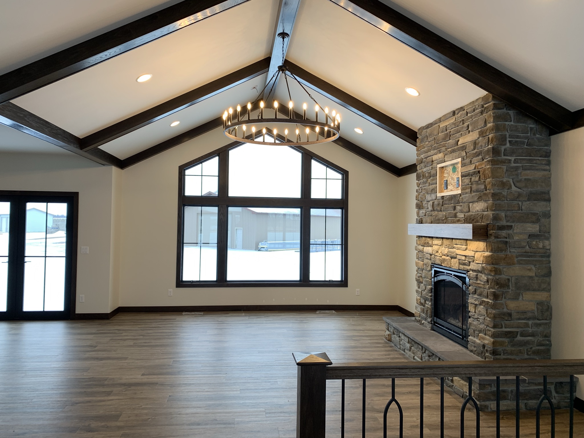 Spacious living room with high vaulted ceilings, exposed dark beams, and large windows. Features a stone fireplace and a rustic chandelier. Cozy ambiance.