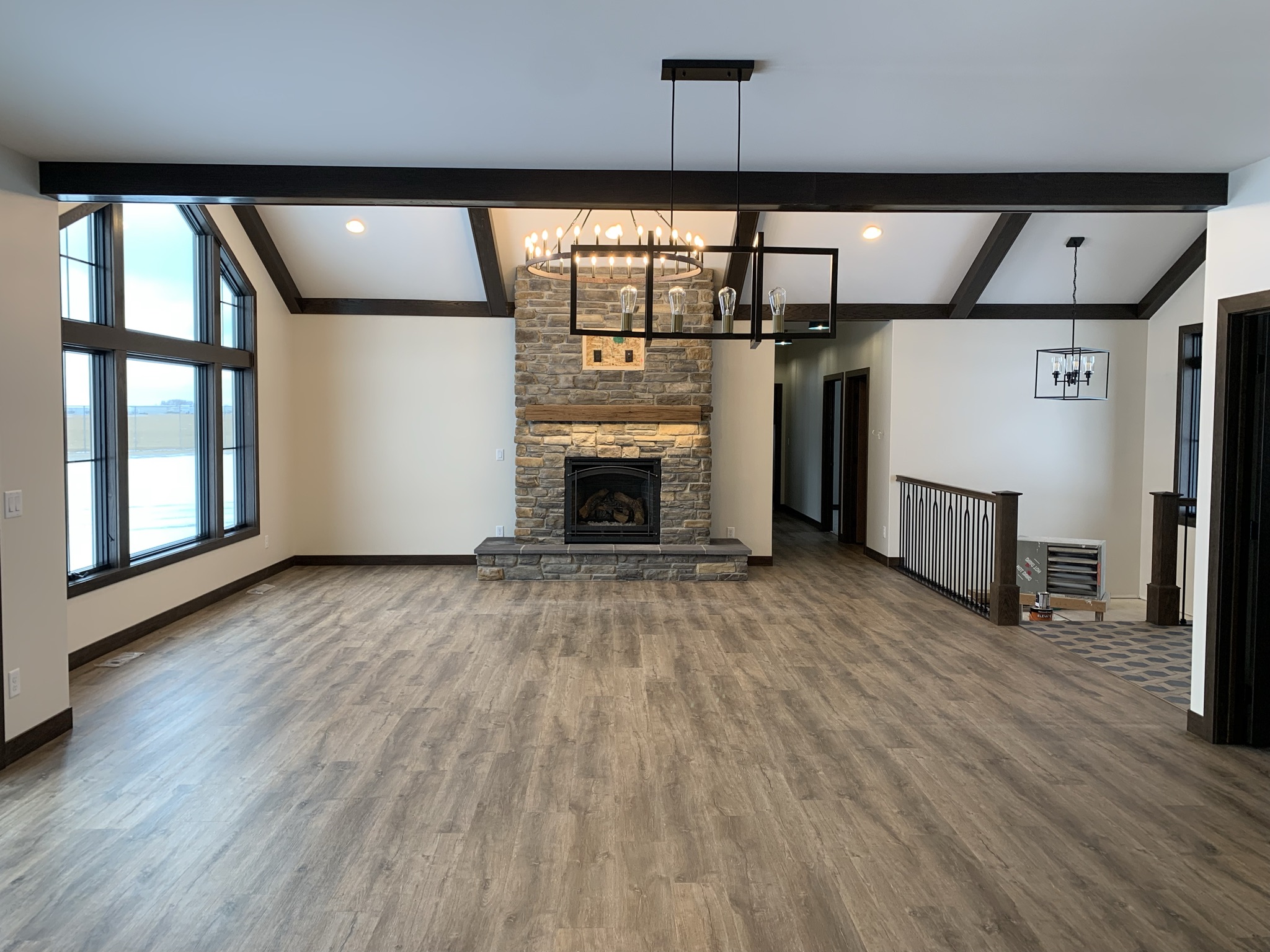 Spacious living room with stone fireplace, wooden beams, and large windows. Modern chandeliers hang from the ceiling, creating a cozy atmosphere.