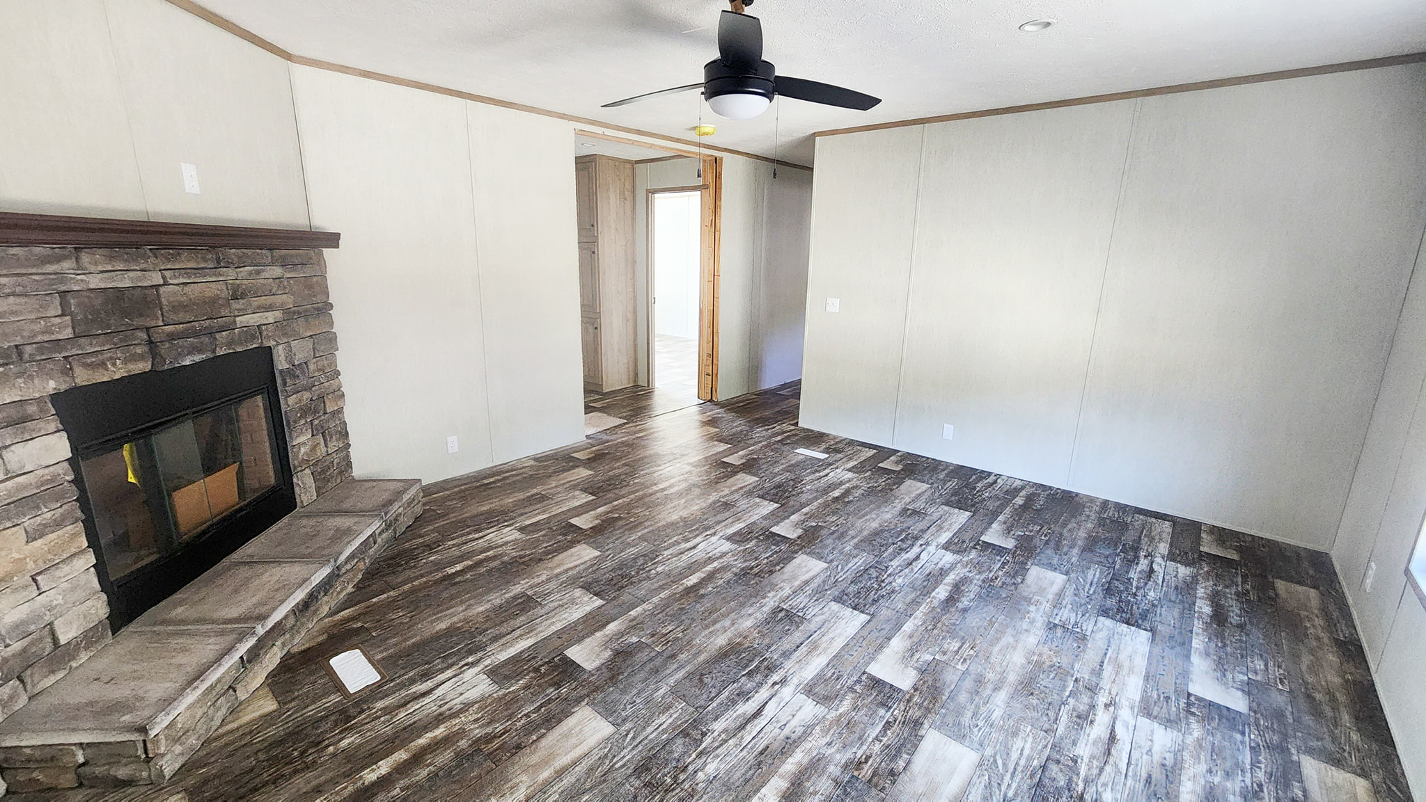 Spacious living room with rustic wood flooring, a stone fireplace on the left, muted beige walls, and a ceiling fan, creating a cozy, airy ambiance.