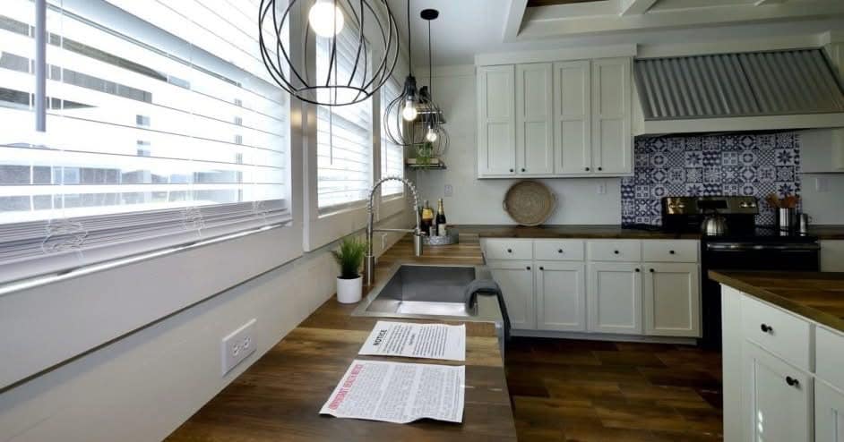 A modern kitchen with wooden countertops, white cabinets, and patterned tile backsplash. Large windows with blinds let in light. A paper is on the counter.