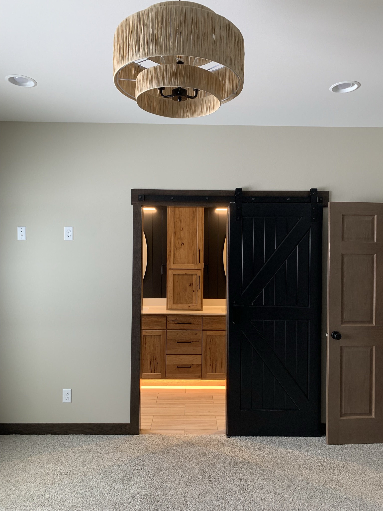 Modern room with beige walls and carpet, featuring a black sliding barn door. Through the door, a lit wooden vanity is visible, with a circular light fixture overhead.