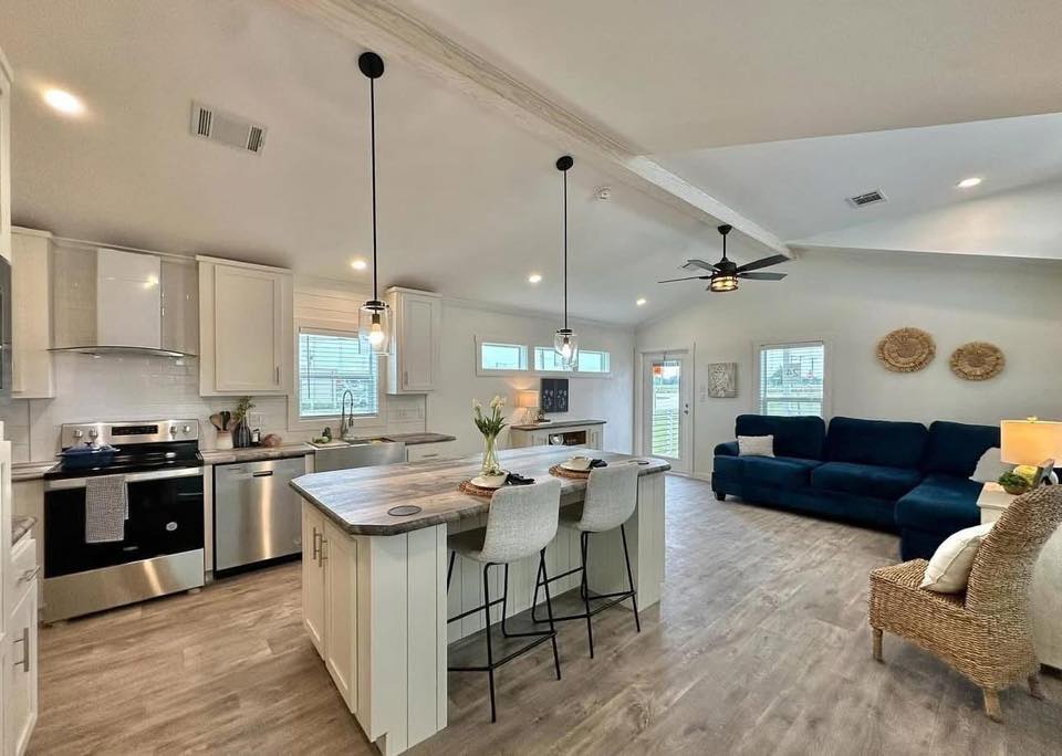 Modern open-plan living space with a kitchen island and bar stools, pendant lighting, and stainless steel appliances. A cozy living area with a navy sectional sofa, wicker chair, and decorative wall baskets creates a welcoming atmosphere.