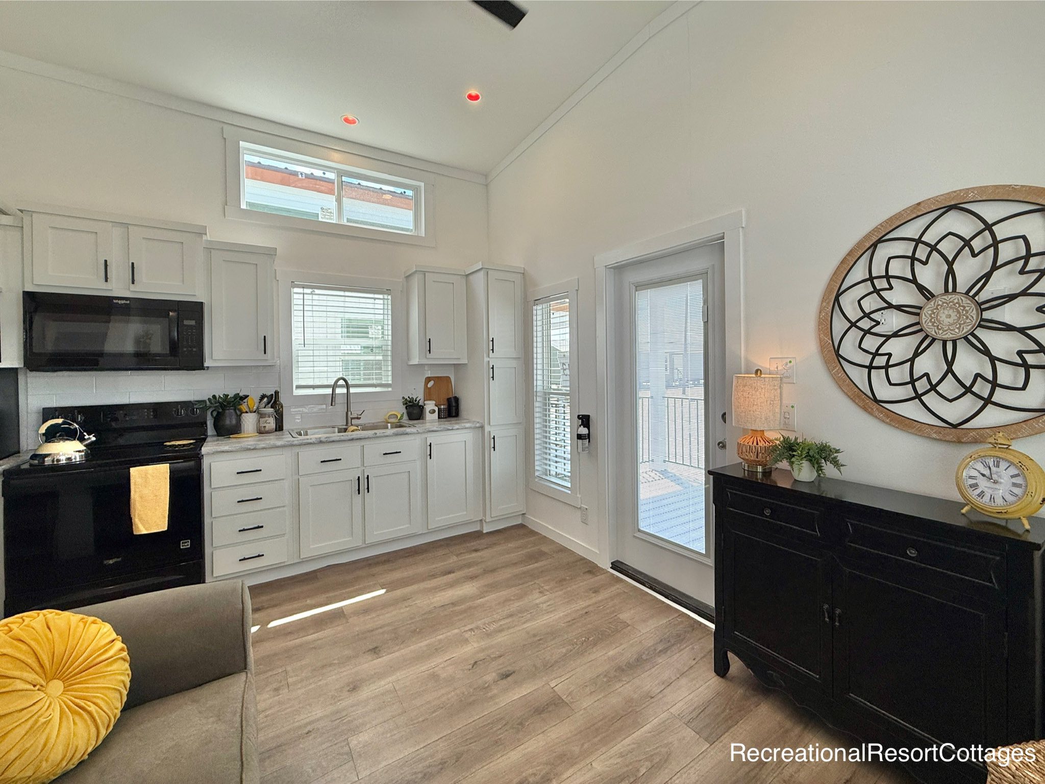 Modern kitchen with white cabinets, black appliances, and wooden flooring. A sunlit window above the sink, decorative clock on the wall, and a cozy ambiance.
