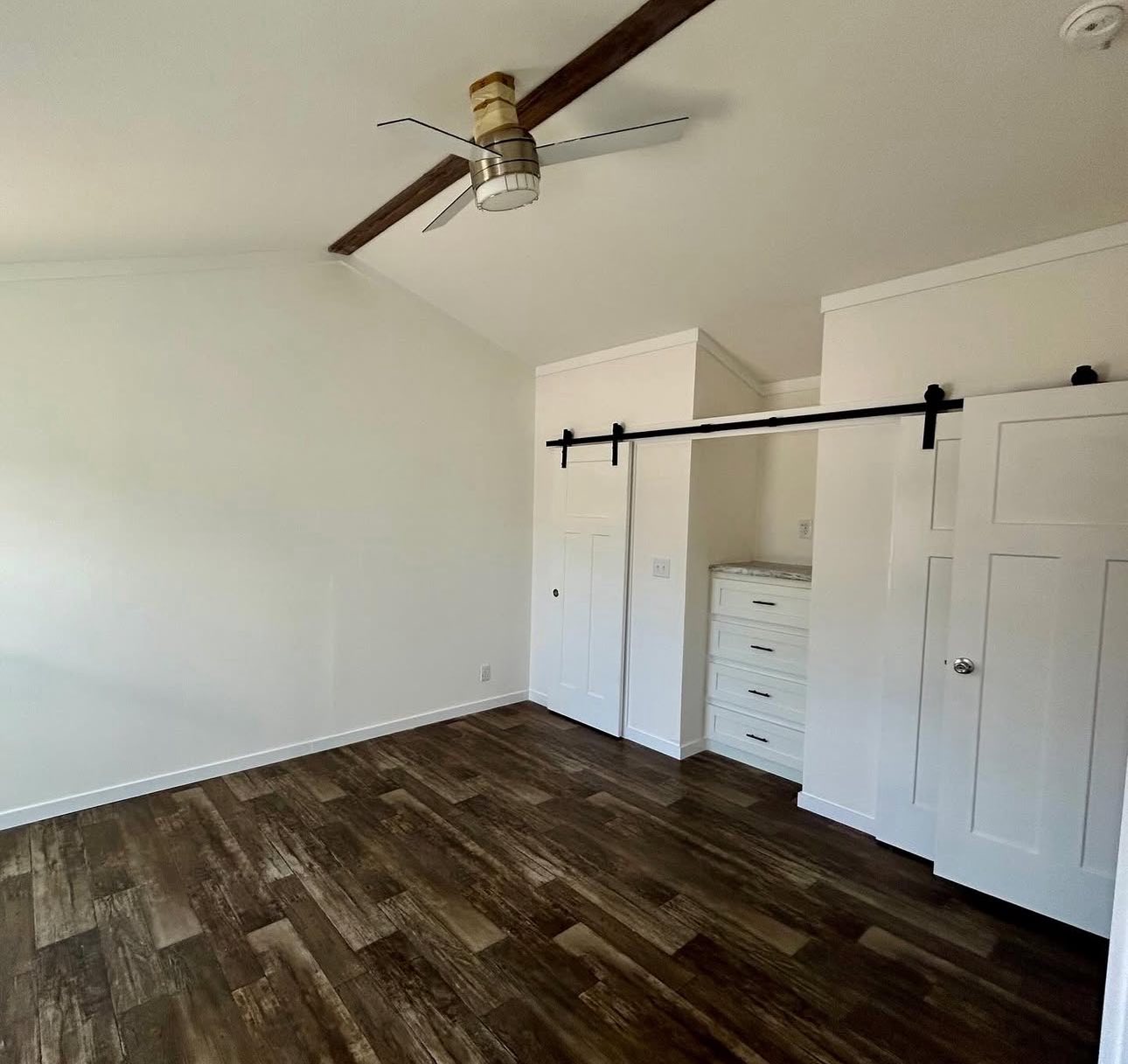 A minimalist empty room with white walls and dark wood flooring. A ceiling fan is mounted on the ceiling. A sliding barn door opens to reveal a small dresser nook.