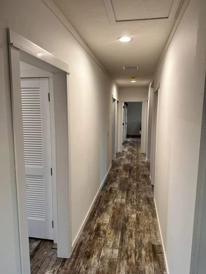 A long, narrow hallway with white walls and wood-patterned laminate flooring. Doors with white frames line both sides, creating a clean, modern look.