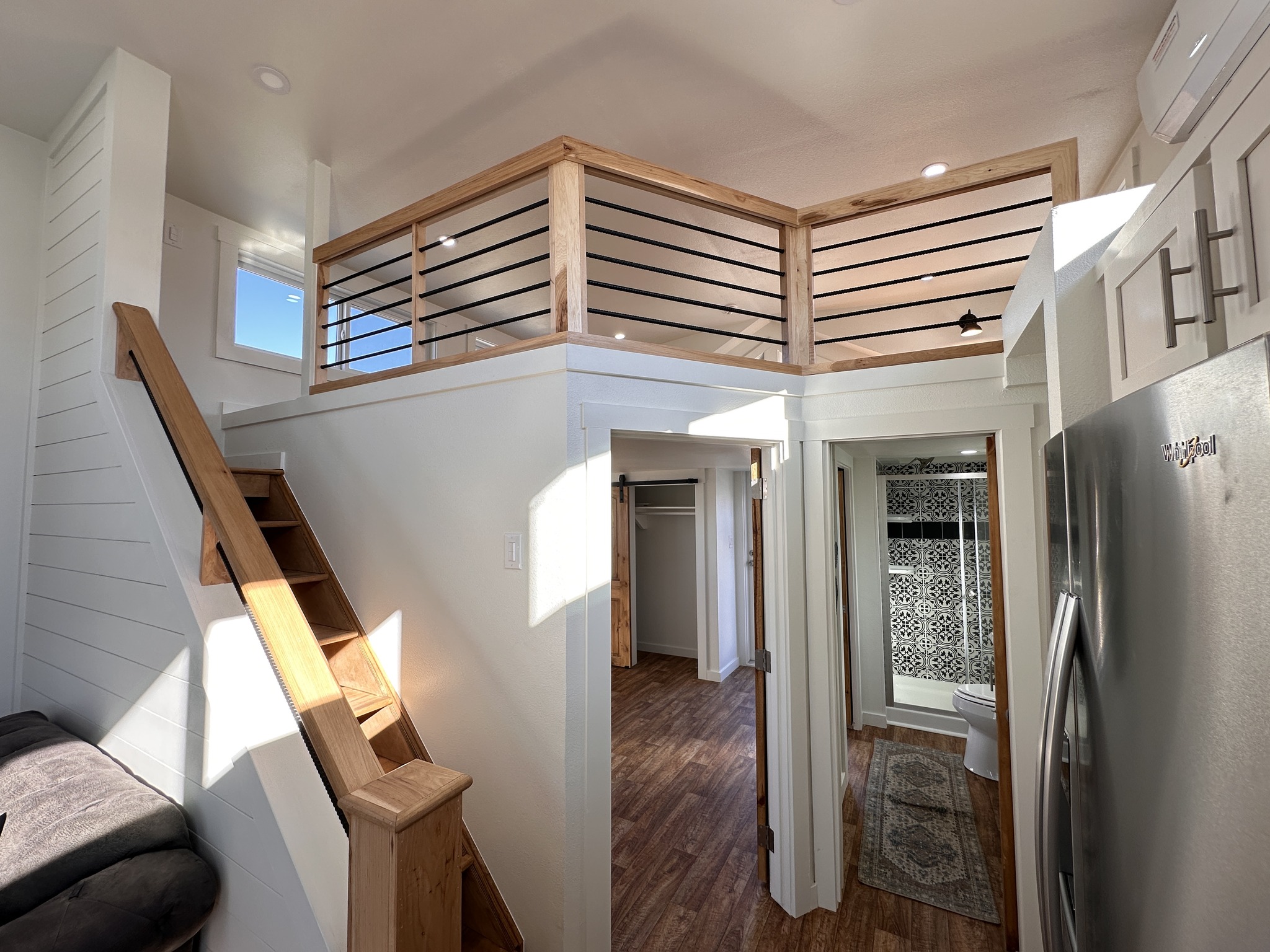 Bright tiny house interior with a wooden loft railing, stairs, and white walls. Open door reveals a patterned bathroom and cozy mood.