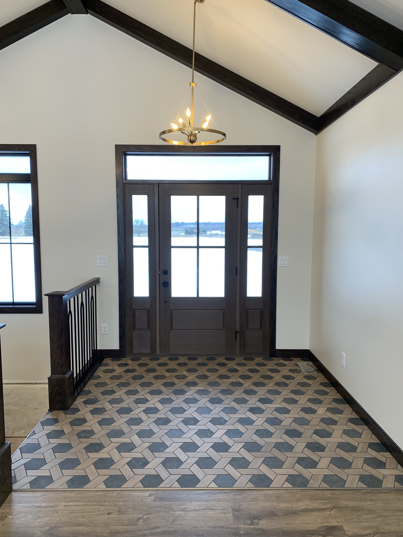 Entrance hallway with a wooden front door featuring glass panels, a geometric patterned tile floor, a modern chandelier, and cream walls.