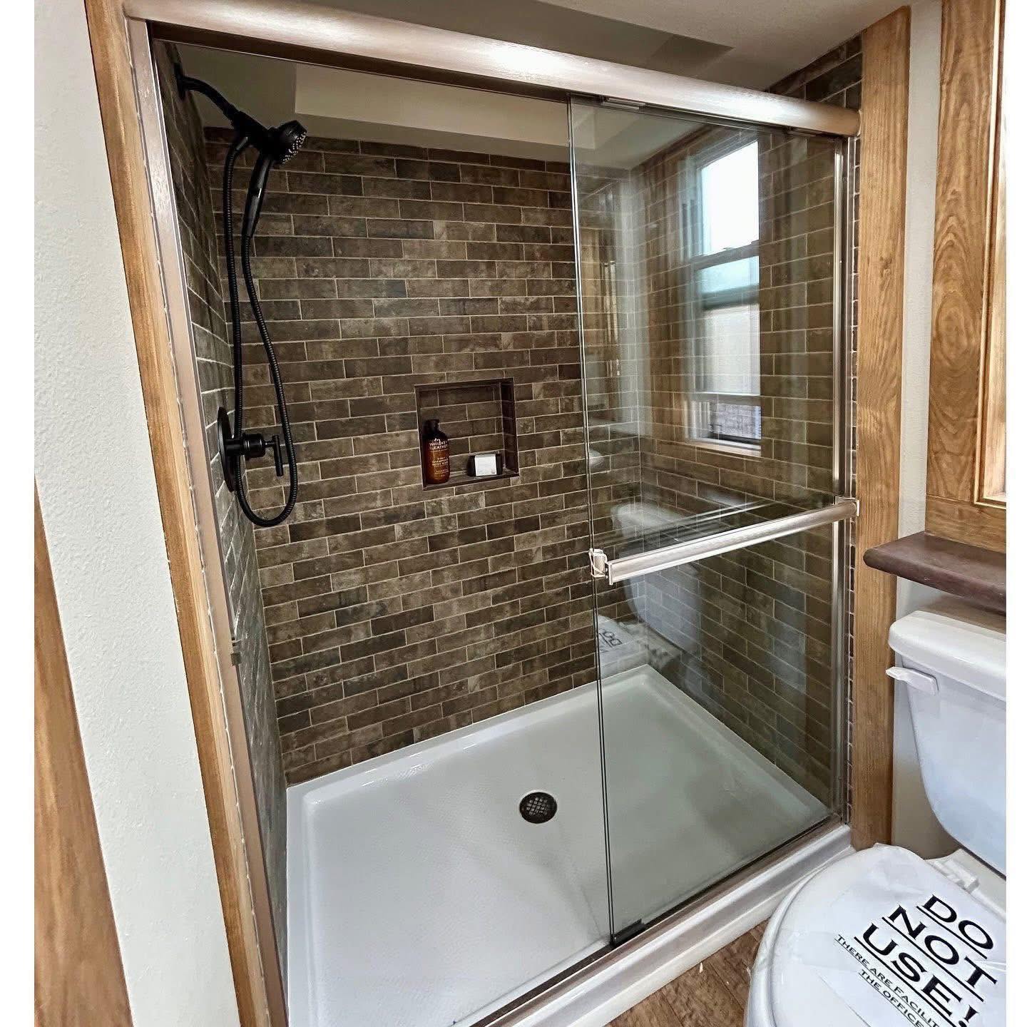 Modern bathroom with a glass shower enclosure and brown tiled walls. A showerhead and toiletries are visible. A "Do Not Use" sign is on the toilet seat.