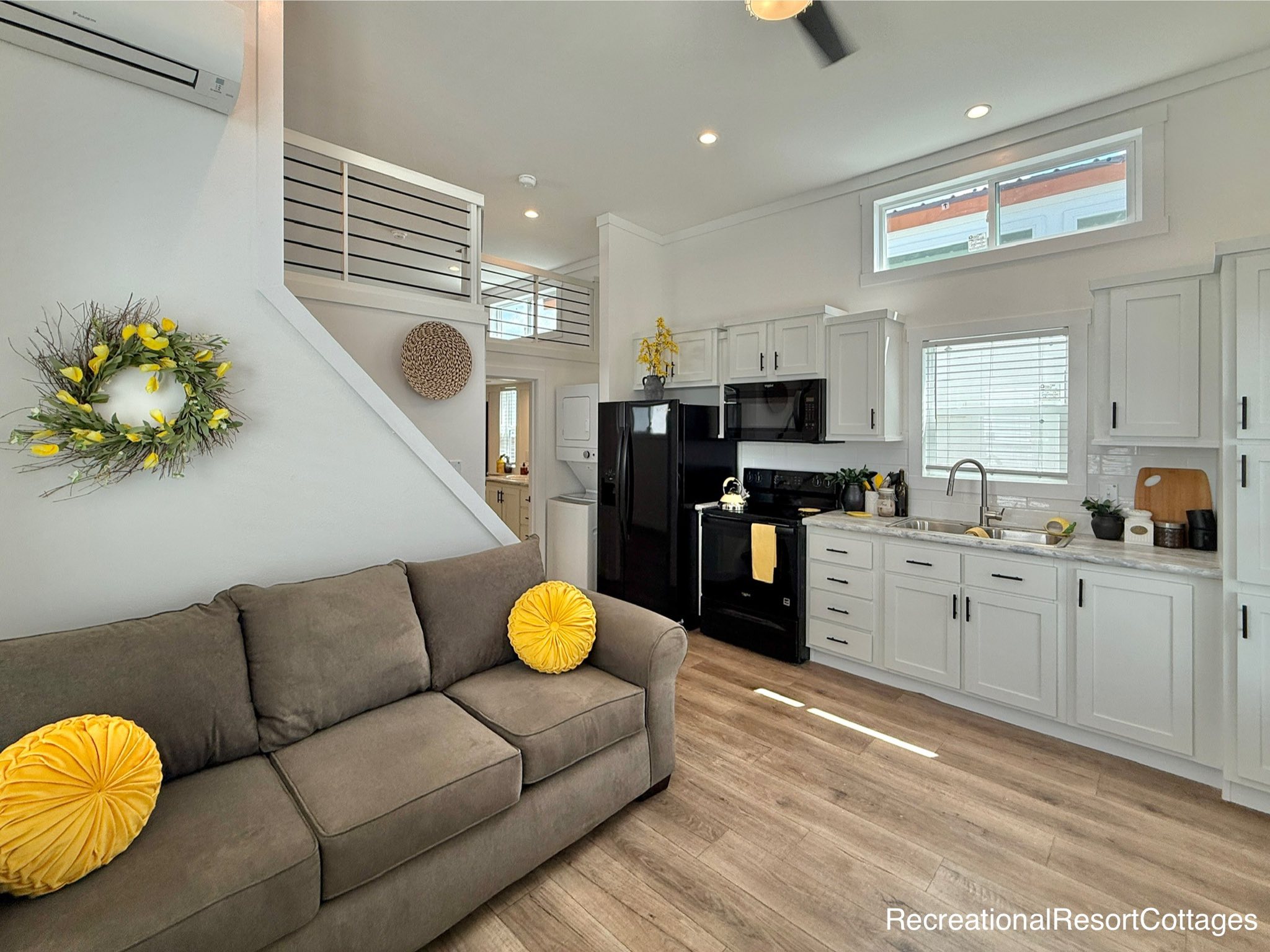 Modern living room and kitchen with high ceilings. A gray sofa has bright yellow cushions. White cabinets, black appliances, and a plant adorn the kitchen.
