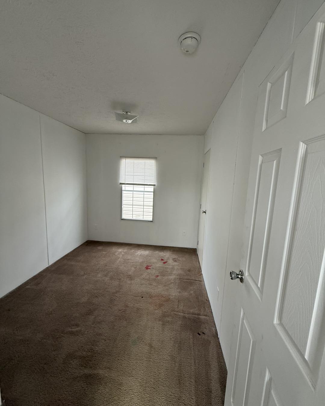 Empty room with white walls and brown carpet, featuring a single window with blinds, a ceiling light, and a smoke detector. The carpet has visible stains.