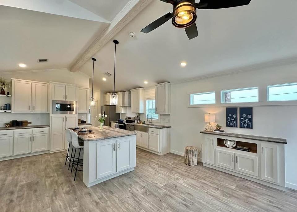 Spacious, modern kitchen with white cabinets, a central island with barstools, pendant lights, and wood flooring. A fan hangs from the ceiling.