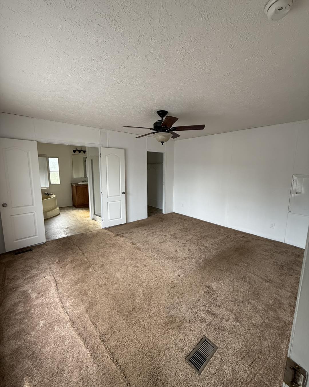 Empty room with beige carpet, white walls, and a ceiling fan. Open doorway reveals a bathroom with a wooden vanity and sink, giving a calm, spacious feel.