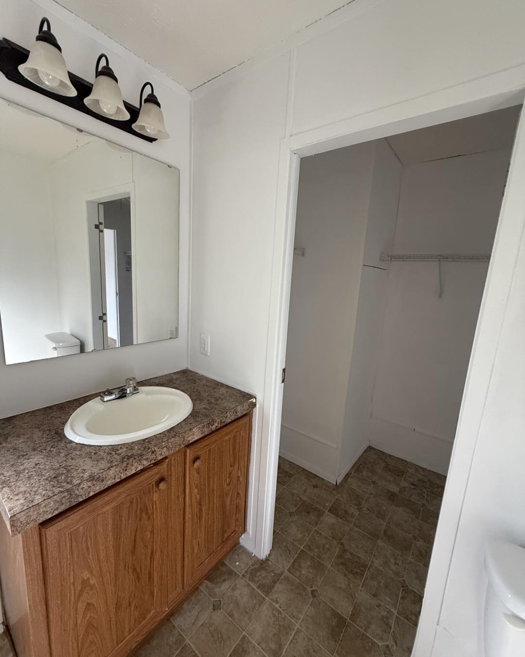 Small bathroom with a wooden vanity and marble countertop. Above the sink is a mirror with three lights. To the right is an empty closet with tiled floors.