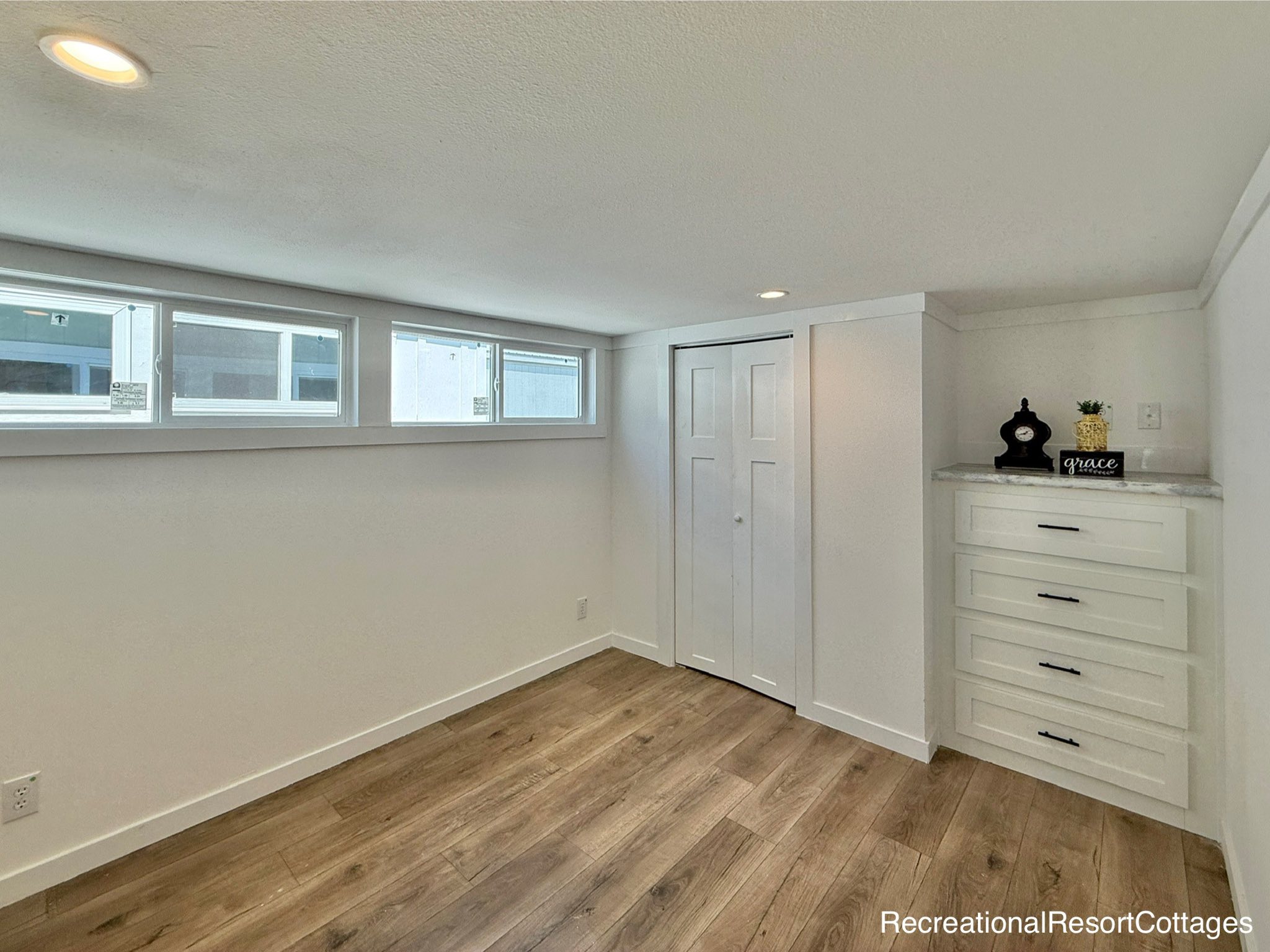 Bright basement with wood flooring, recessed lighting, and small windows. Features a white dresser and closet, creating a clean, cozy atmosphere.