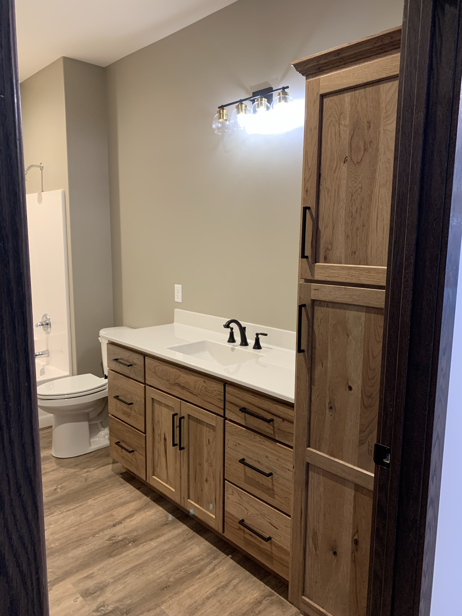 Modern bathroom with light wood cabinets and drawers, white sink with black fixtures, and lit mirror. Toilet and bathtub in background on wood floor. Cozy ambiance.