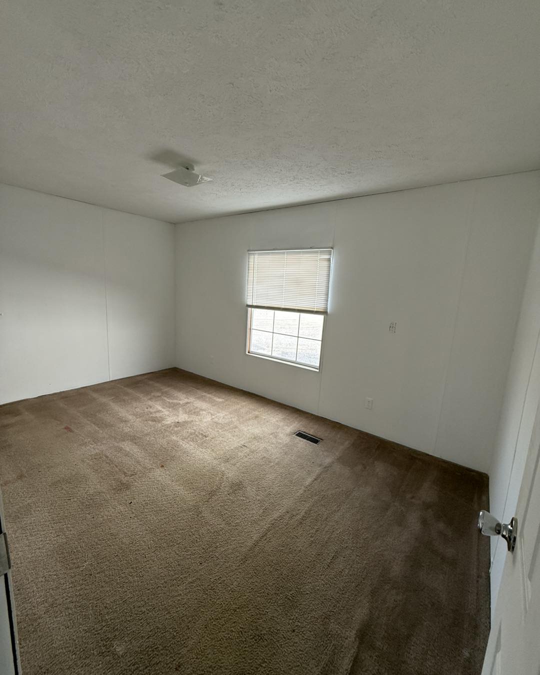 An empty room with white walls and a brown carpet. A single window with closed blinds lets in some light, creating a calm, neutral atmosphere.