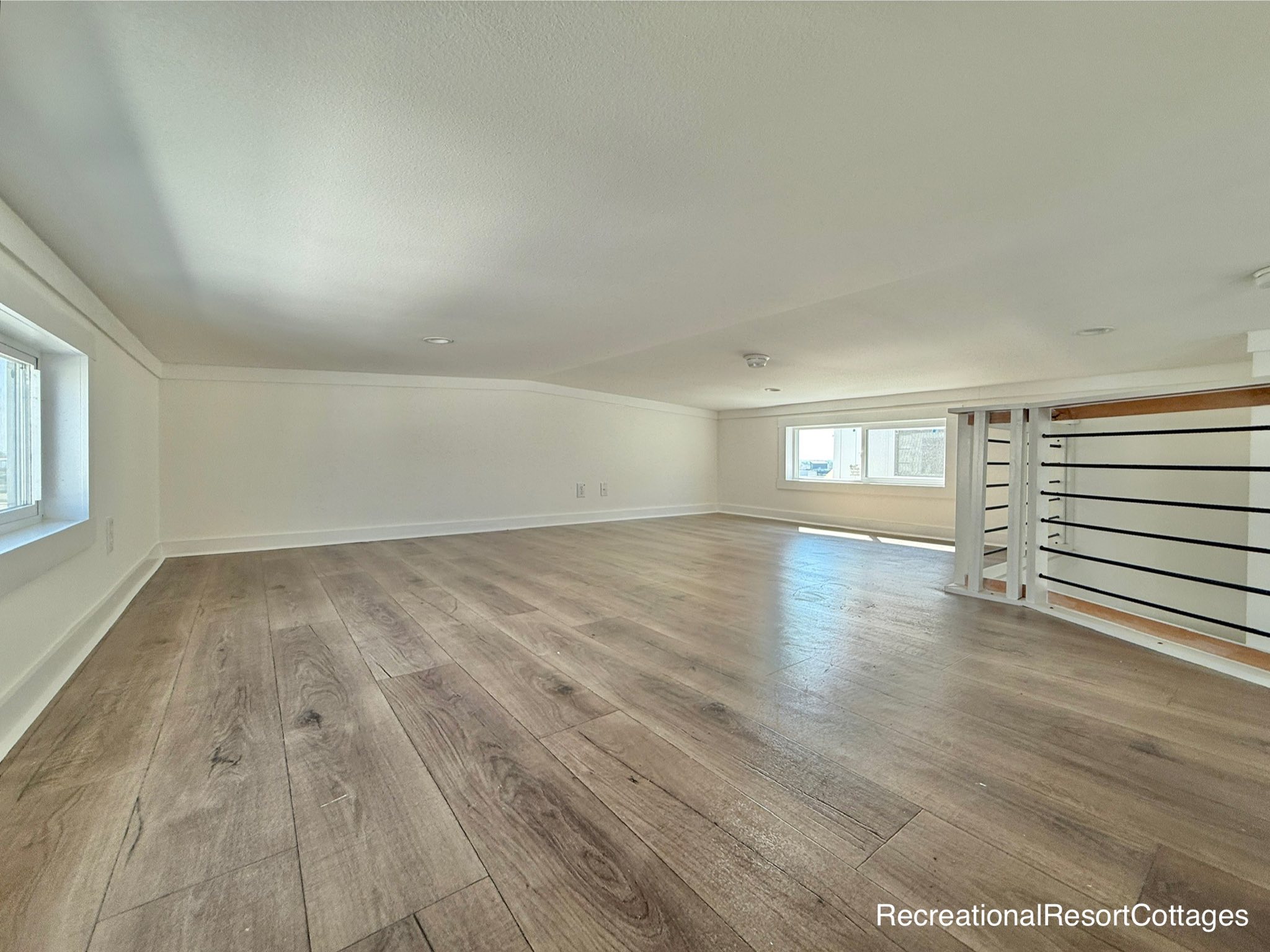 Spacious attic with light wood flooring, white walls, and low ceiling. Two windows provide natural light. A minimalist railing adds a modern touch.