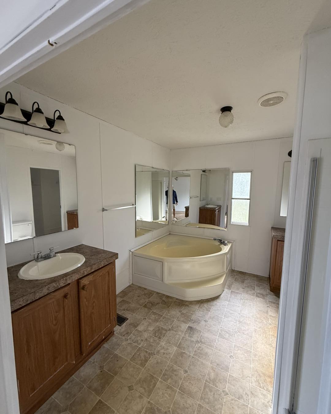 A spacious bathroom with beige tiles features a corner bathtub, wooden cabinets, a countertop sink, mirrored walls, and ample natural light.